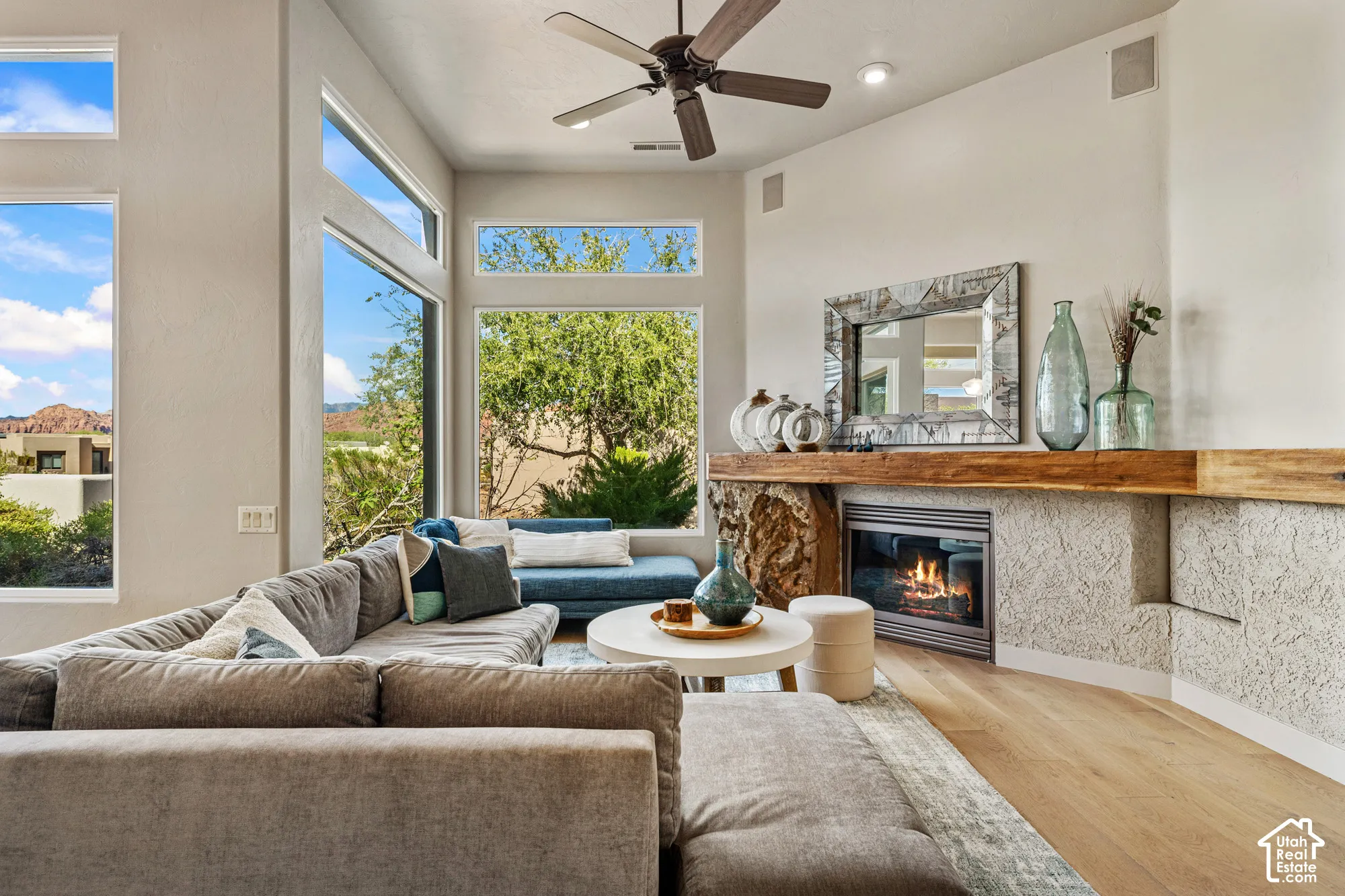 Vaulted ceiling with an integrated ceiling fan, large picture windows, wood-finish flooring, built-in fireplace, and a stone-clad mantle with a wood beam