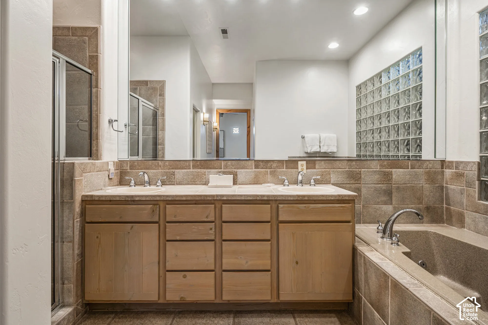 Dual sink vanity with light wood cabinetry, a built-in tiled bathtub, a glass block window, a tiled shower, and a large wall mirror