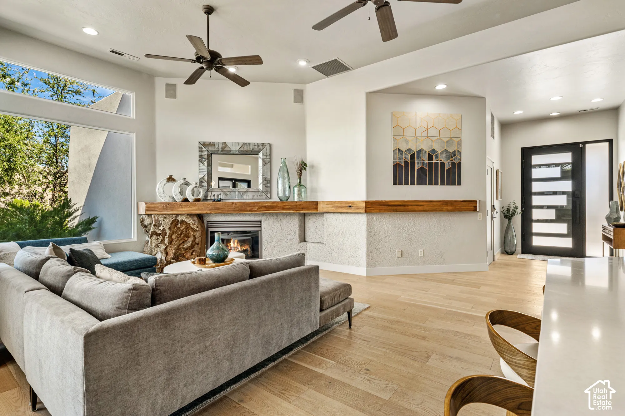 Light wood-finish flooring, stone-clad fireplace with wood mantle, modern dark-frame entry door with frosted glass, recessed lighting, and ceiling fans