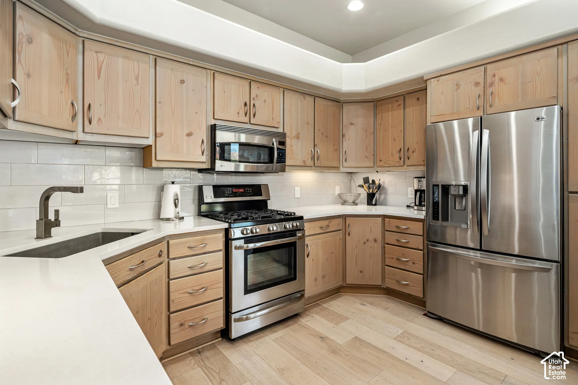 Light-toned wood cabinetry, stainless steel appliances, white subway tile backsplash, solid surface countertops, and light wood-finish flooring