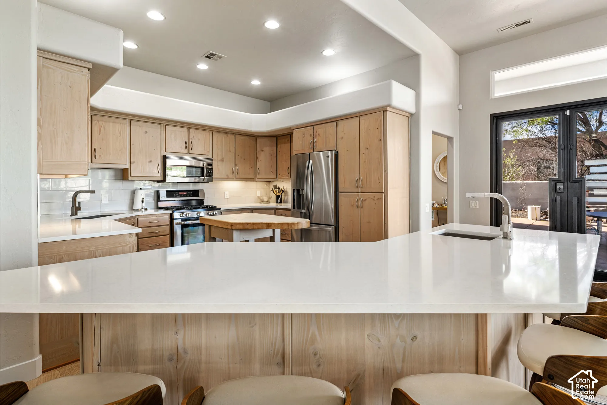Kitchen featuring light wood cabinetry, stainless steel appliances, a white subway tile backsplash, and a large white countertop with an integrated sink