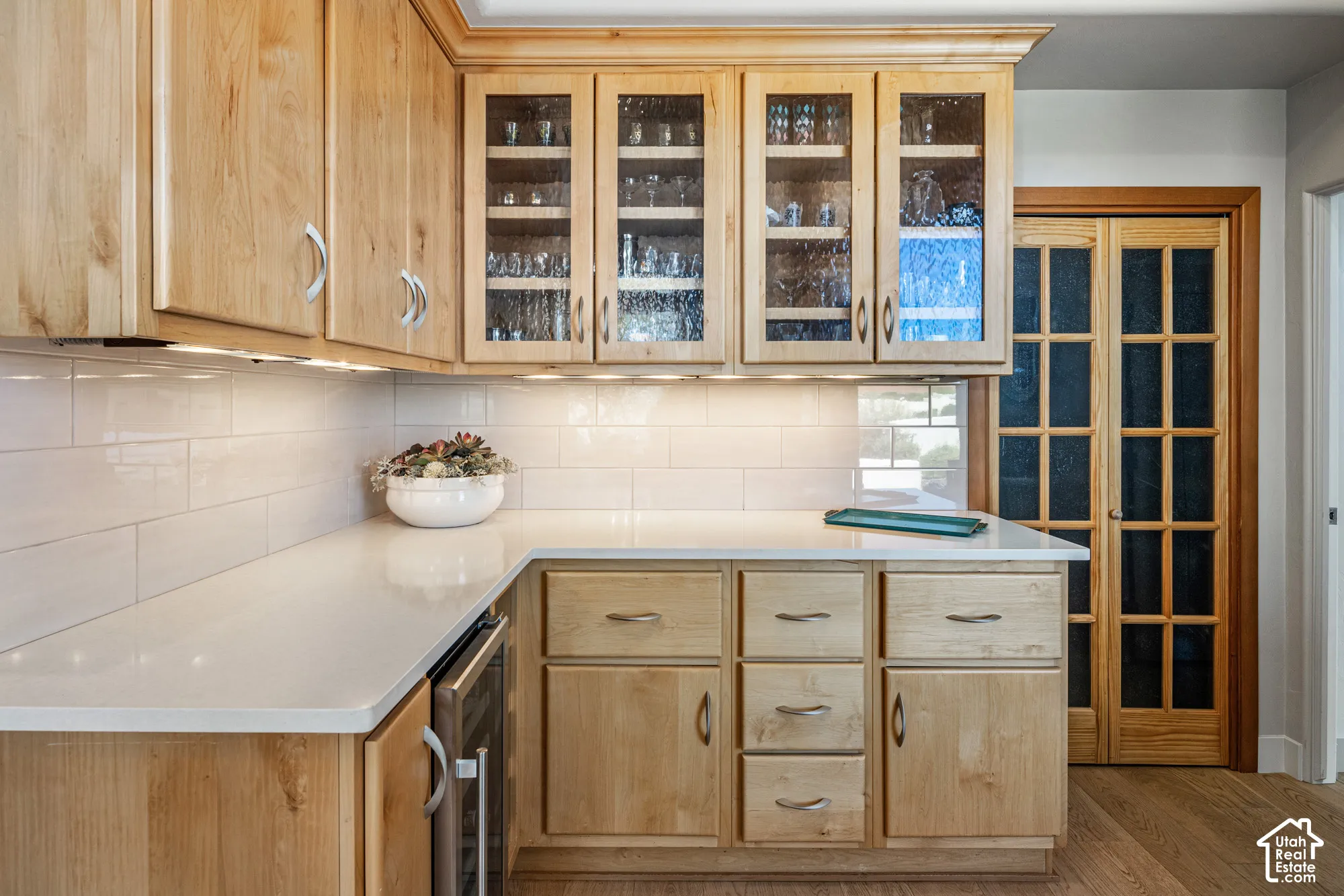 Wood-finish cabinetry with glass-front uppers, solid surface countertops, white subway tile backsplash, under-cabinet lighting, and wood-finish flooring