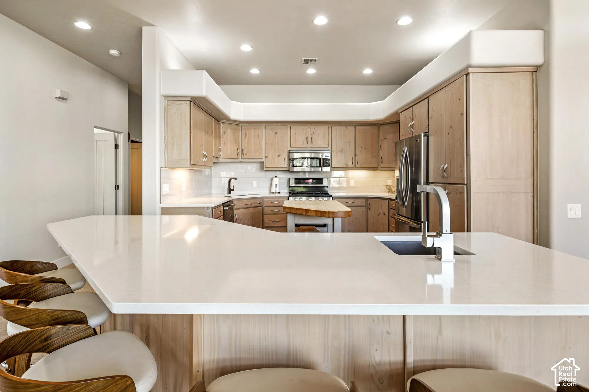 Kitchen featuring recessed lighting, a built-in stainless steel refrigerator, light wood-finish cabinetry, a large kitchen island with an undermount sink, and white countertops