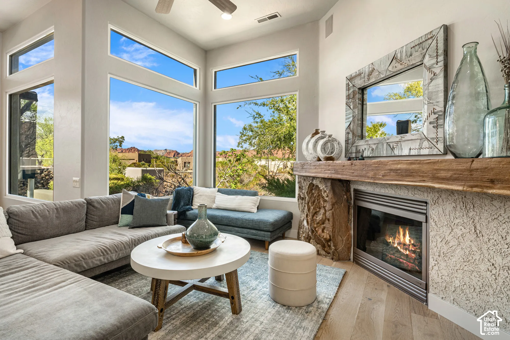 Expansive window array, wood-finish flooring, recessed lighting, ceiling fan, and a fireplace with a rough-hewn stone mantel