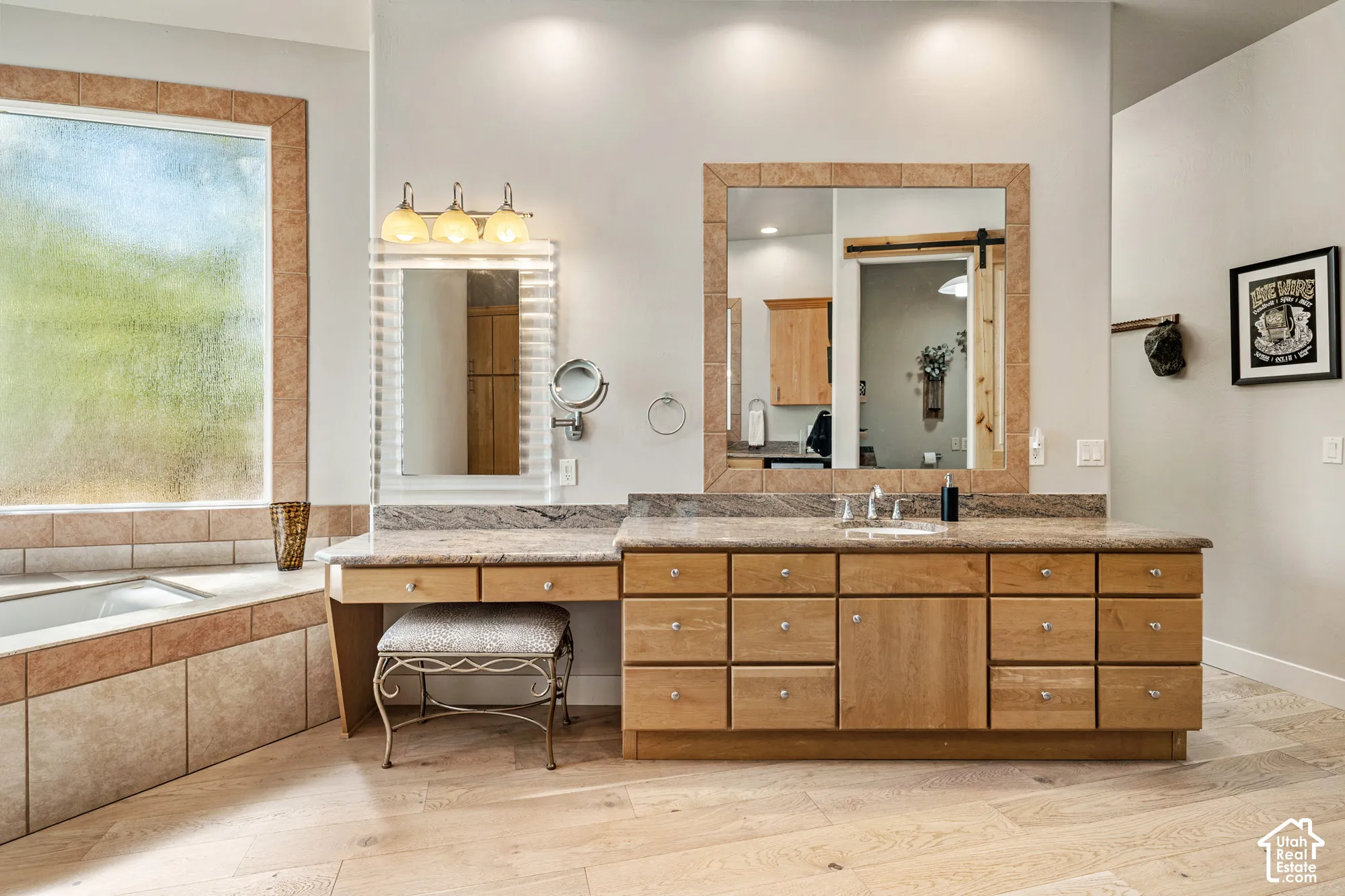 Spacious bathroom featuring a built-in soaking tub with tile surround, expansive wood-finish vanity with dual sinks, and light wood-finish flooring