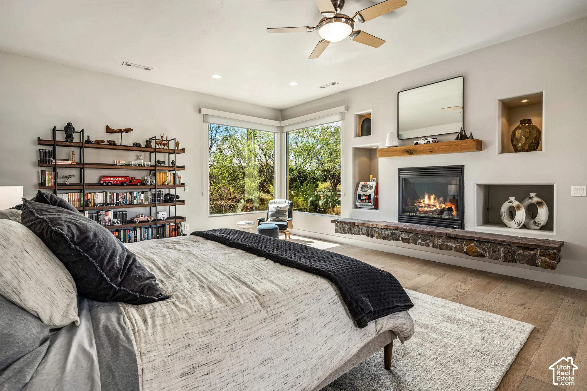 Corner windows, wood-finish flooring, built-in shelving, ceiling fan, and a fireplace with a stone-clad hearth