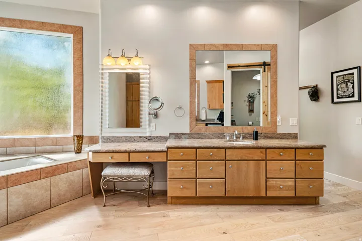Spacious bathroom featuring a built-in soaking tub with tile surround, expansive wood-finish vanity with dual sinks, and light wood-finish flooring