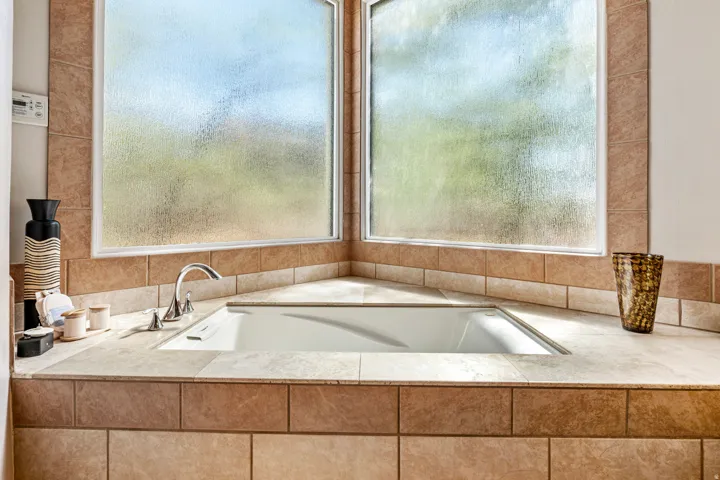 Built-in corner bathtub with a polished chrome faucet, surrounded by tiled decking and two large privacy glass windows