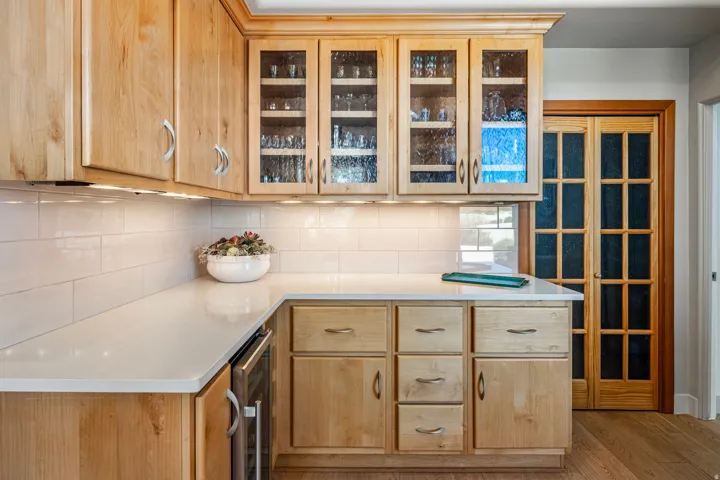 Wood-finish cabinetry with glass-front uppers, solid surface countertops, white subway tile backsplash, under-cabinet lighting, and wood-finish flooring