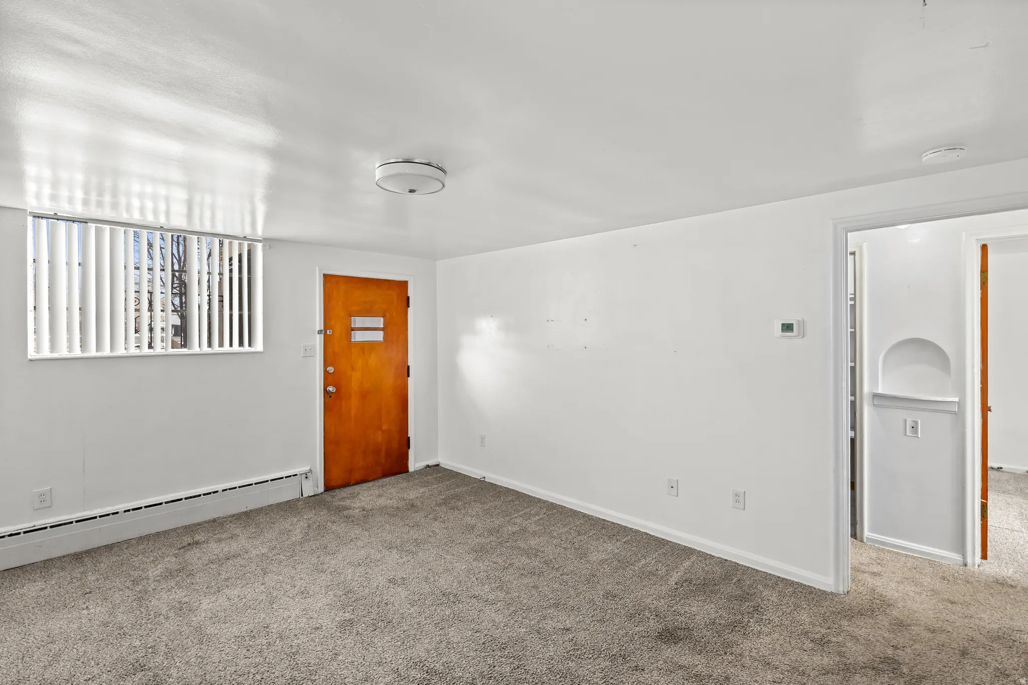 Carpeted interior with white walls, window with vertical blinds, wood-finish door, and flush-mount ceiling light