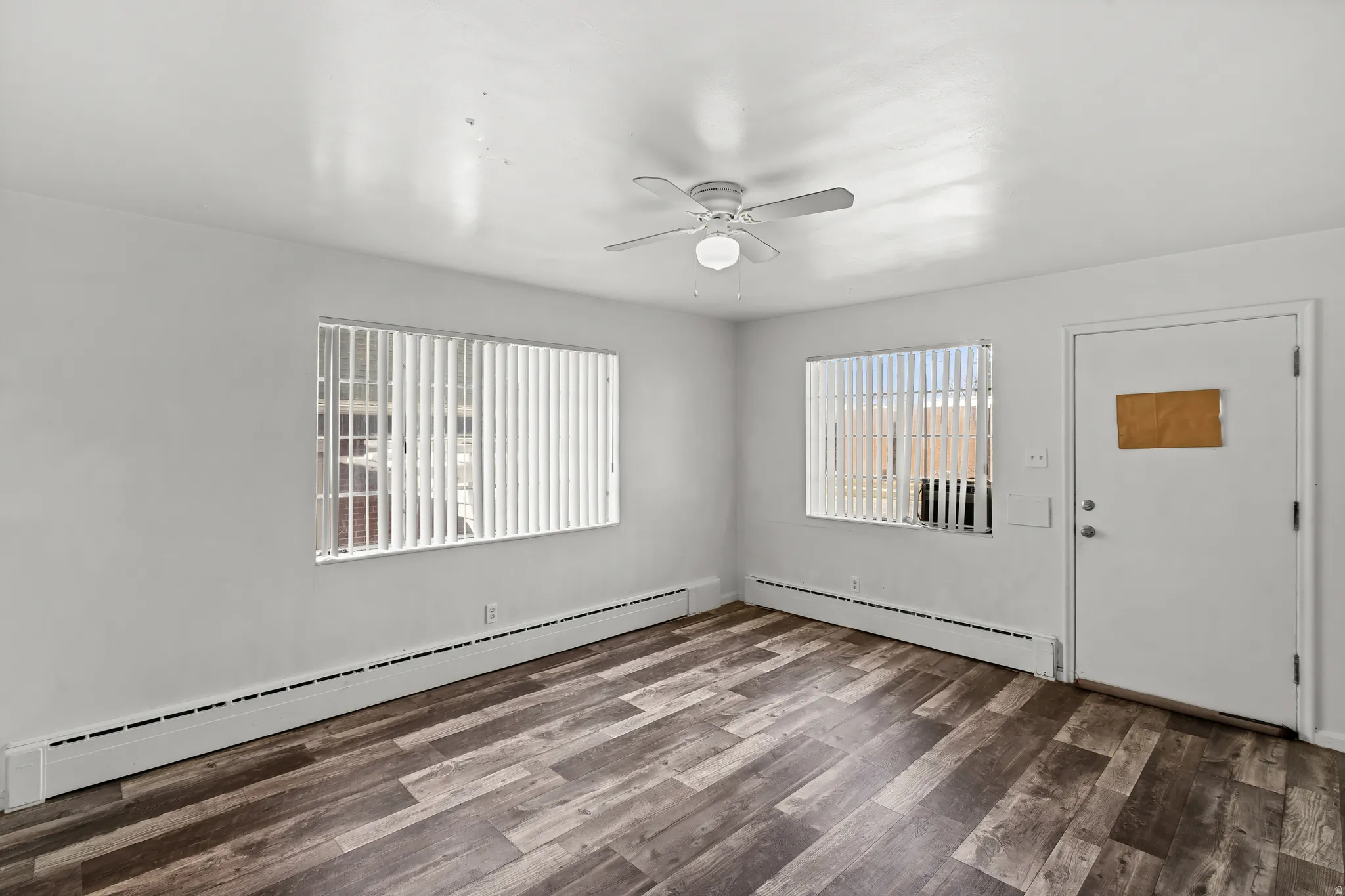 Wood-finish flooring, white walls, two windows with vertical blinds, and a ceiling fan with integrated lighting