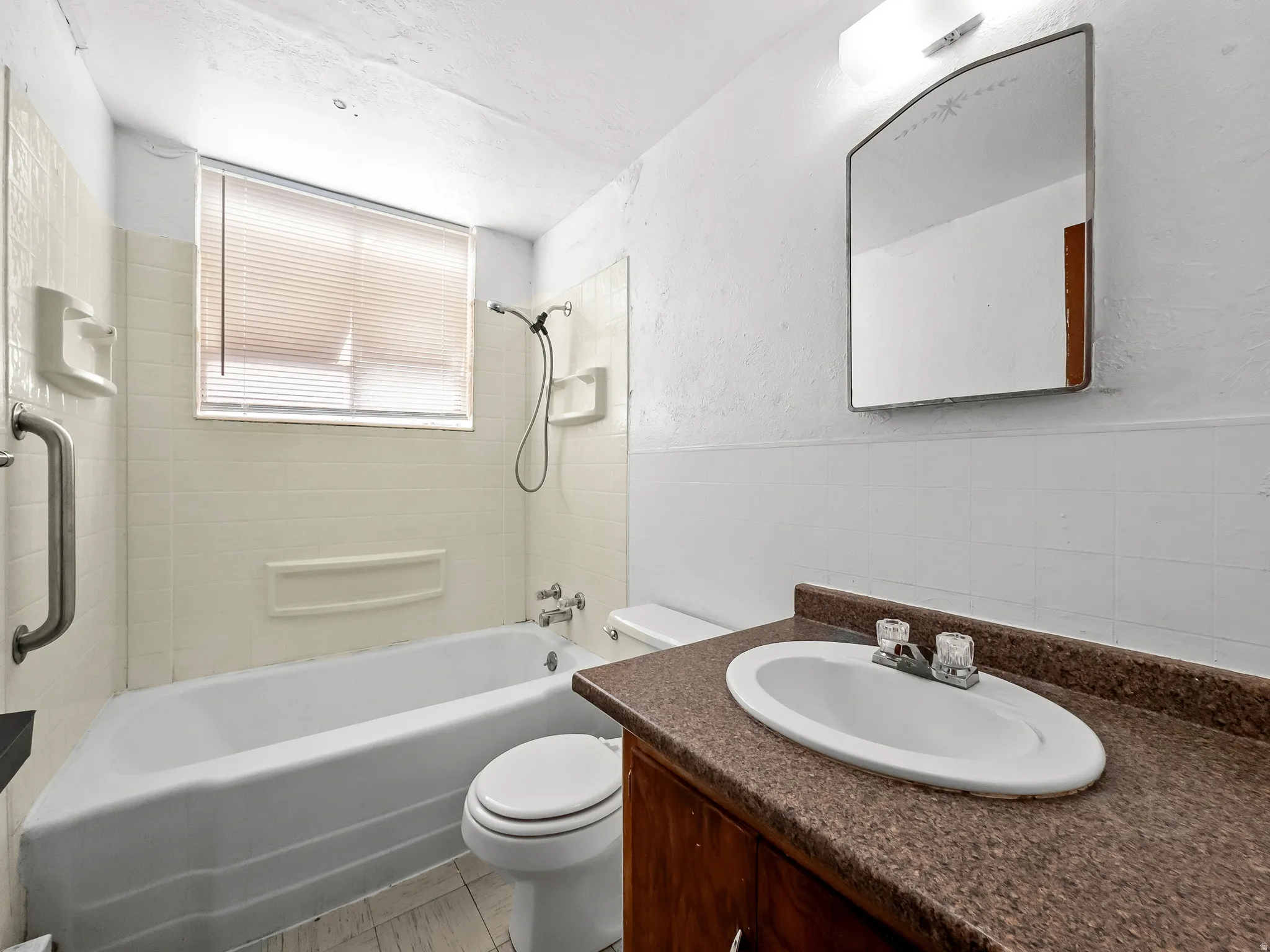 White wall tiling, built-in bathtub, vanity with integrated sink, wall-mounted mirror, and window with blinds