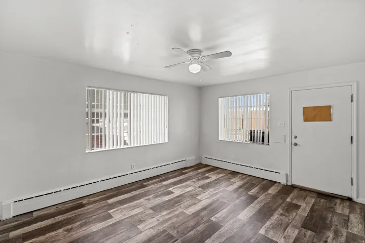Wood-finish flooring, white walls, two windows with vertical blinds, and a ceiling fan with integrated lighting