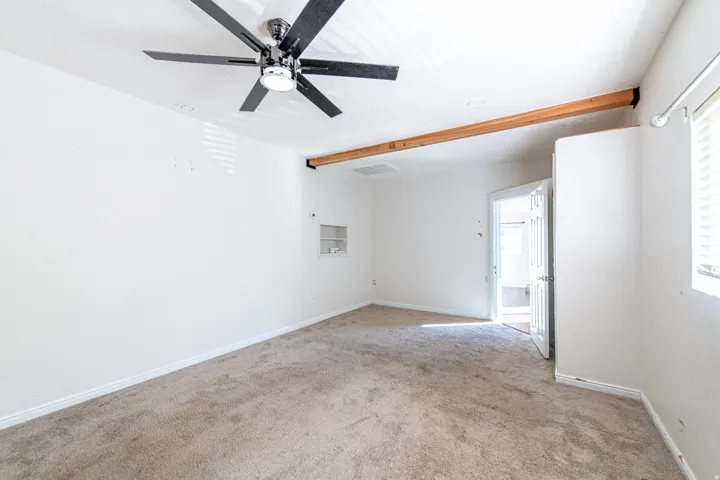 Wall-mounted ceiling fan, exposed wood beam, built-in wall niche, window with blinds, and neutral carpeting