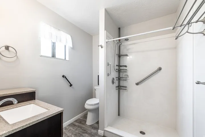 White shower stall with grab bars and built-in shelving, vanity with integrated sink and stone-finish countertop, wood-finish flooring, and a window
