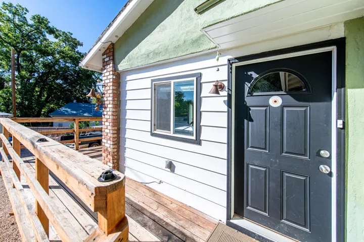 White siding exterior with brick chimney accent, wood railing, and a black paneled door with an arched glass insert