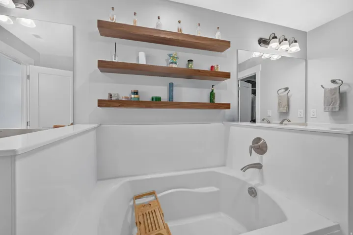 Built-in bathtub with brushed nickel fixtures, floating wood-finish shelving, and expansive vanity mirrors