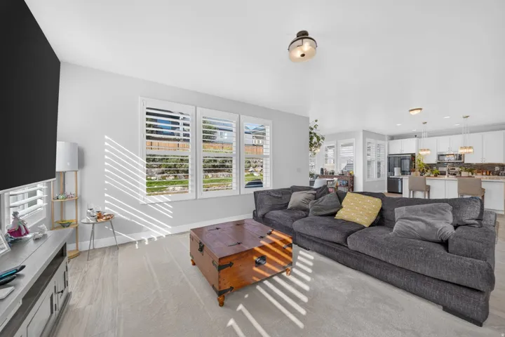 Spacious open concept living area featuring light-toned flooring, three large windows with white louvered shutters, and recessed ceiling lighting