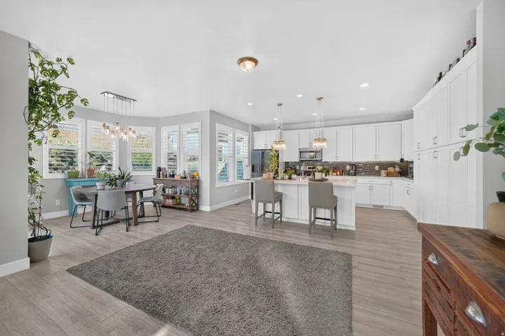 Open-concept living space with wood-finish flooring, a dining area featuring a bay window with plantation shutters, and a kitchen with white cabinetry, a central island, and pendant lighting