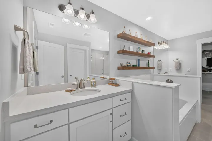 Vanity with white cabinetry and light-toned countertop, undermount sink with brushed nickel faucet, large wall-mounted mirror, built-in bathtub, and floating wood shelves