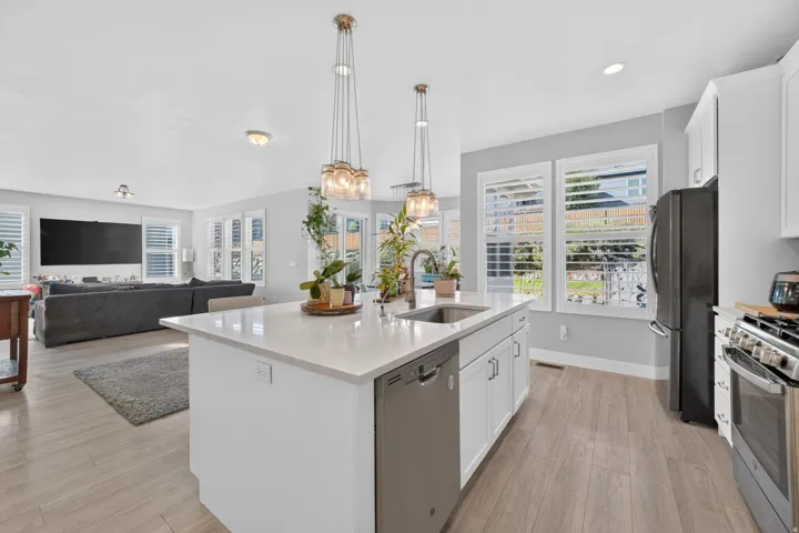 Modern kitchen featuring a large white island with waterfall countertop edge, stainless steel appliances including a built-in dishwasher and double wall ovens, white cabinetry with hardware accents, pendant lighting above the island, and wood-finish floor