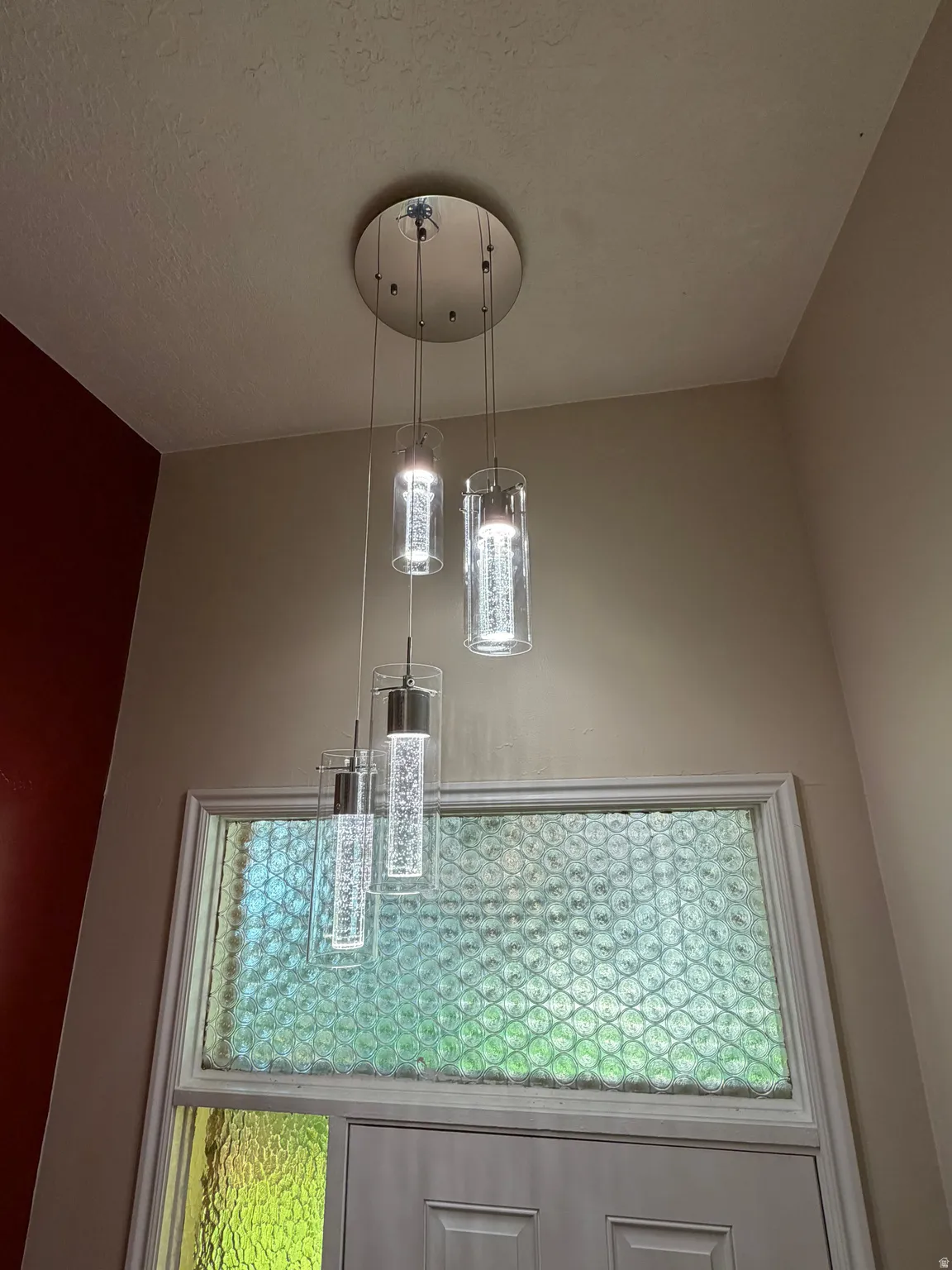 Modern pendant lighting fixture featuring three cylindrical glass shades suspended from a brushed nickel canopy, illuminating the entryway above a solid-core door with decorative glass panel insert