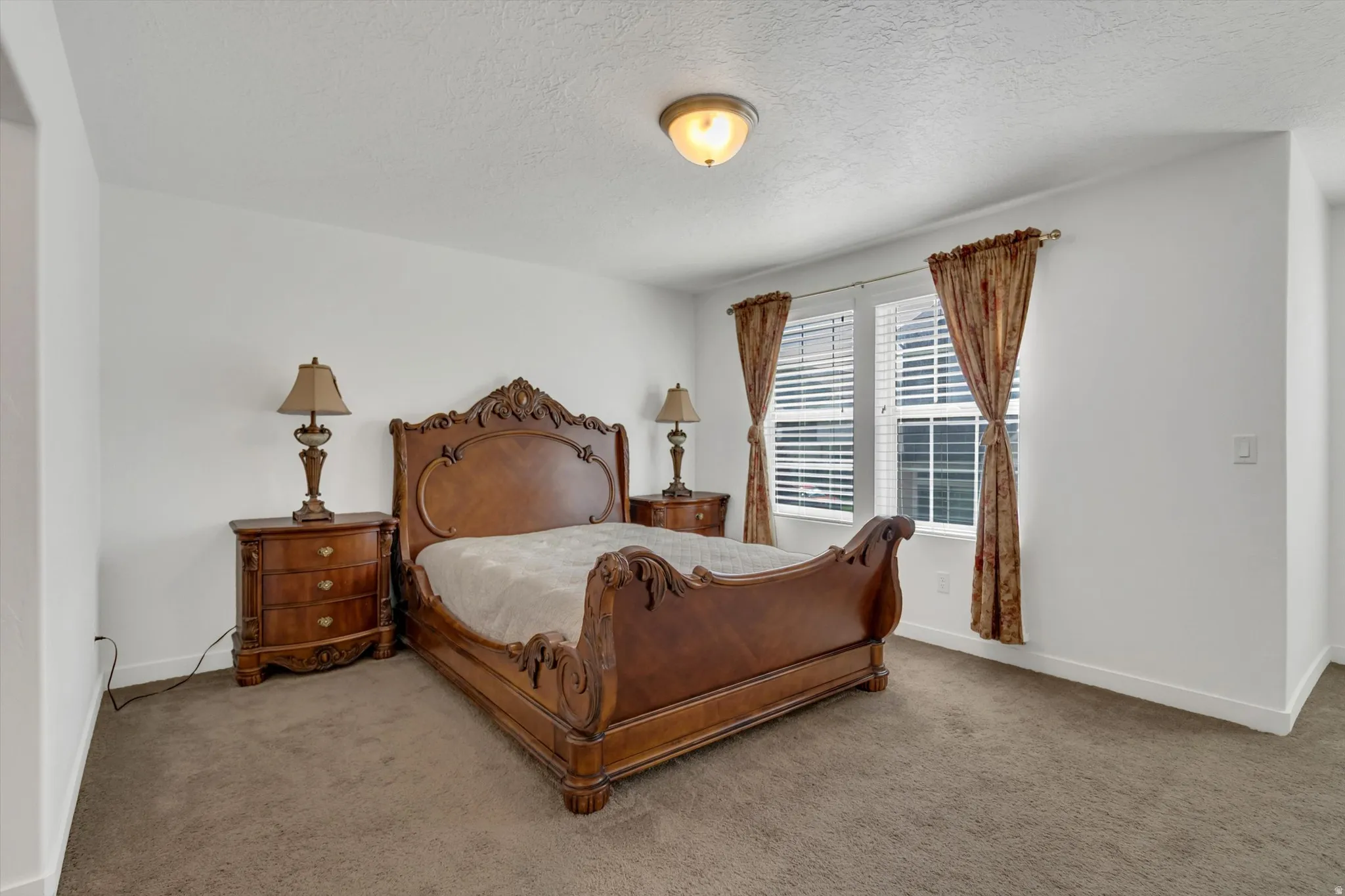 Spacious primary bedroom featuring ornate wood-carved bed frame, coordinating nightstands with brass table lamps, neutral carpet flooring, and large multi-pane windows dressed with bronze-toned drapery