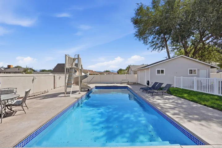 Rectangular swimming pool with a slide, surrounded by a concrete pool deck, fenced by a privacy wall, and complemented by a separate ancillary structure