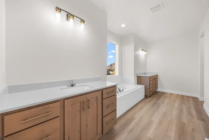 Dual wood-finish vanities with light-toned countertops, integrated soaking tub, wood-finish flooring, and contemporary wall sconces