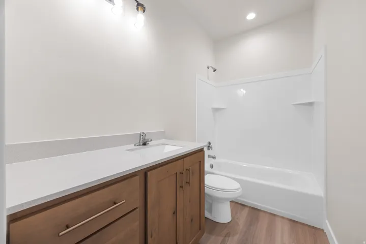 Wood-finish flooring, light-toned walls, vanity with wood-finish cabinetry, light-toned countertop, and a built-in bathtub with shower