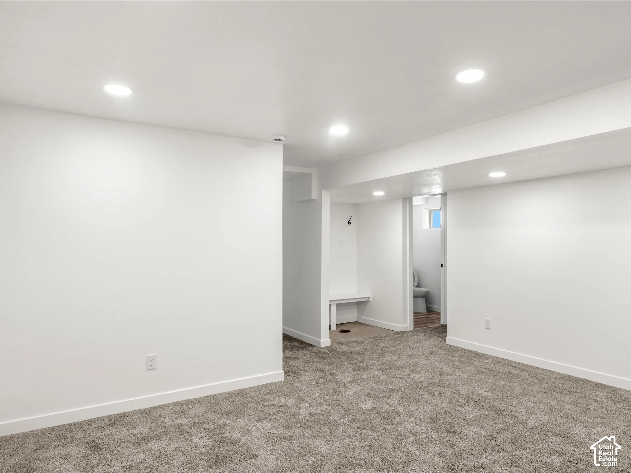 Recessed ceiling lighting, white walls, gray carpet flooring, built-in storage bench, and a partial bathroom with a toilet