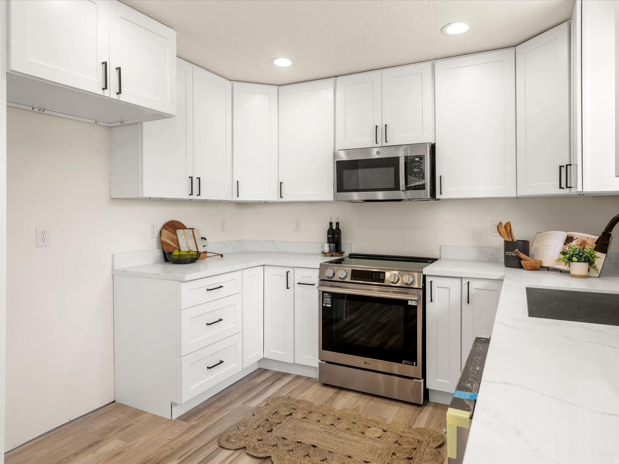 White cabinetry with black hardware, stainless steel appliances, light-toned wood-finish flooring, recessed lighting, and light-toned countertops