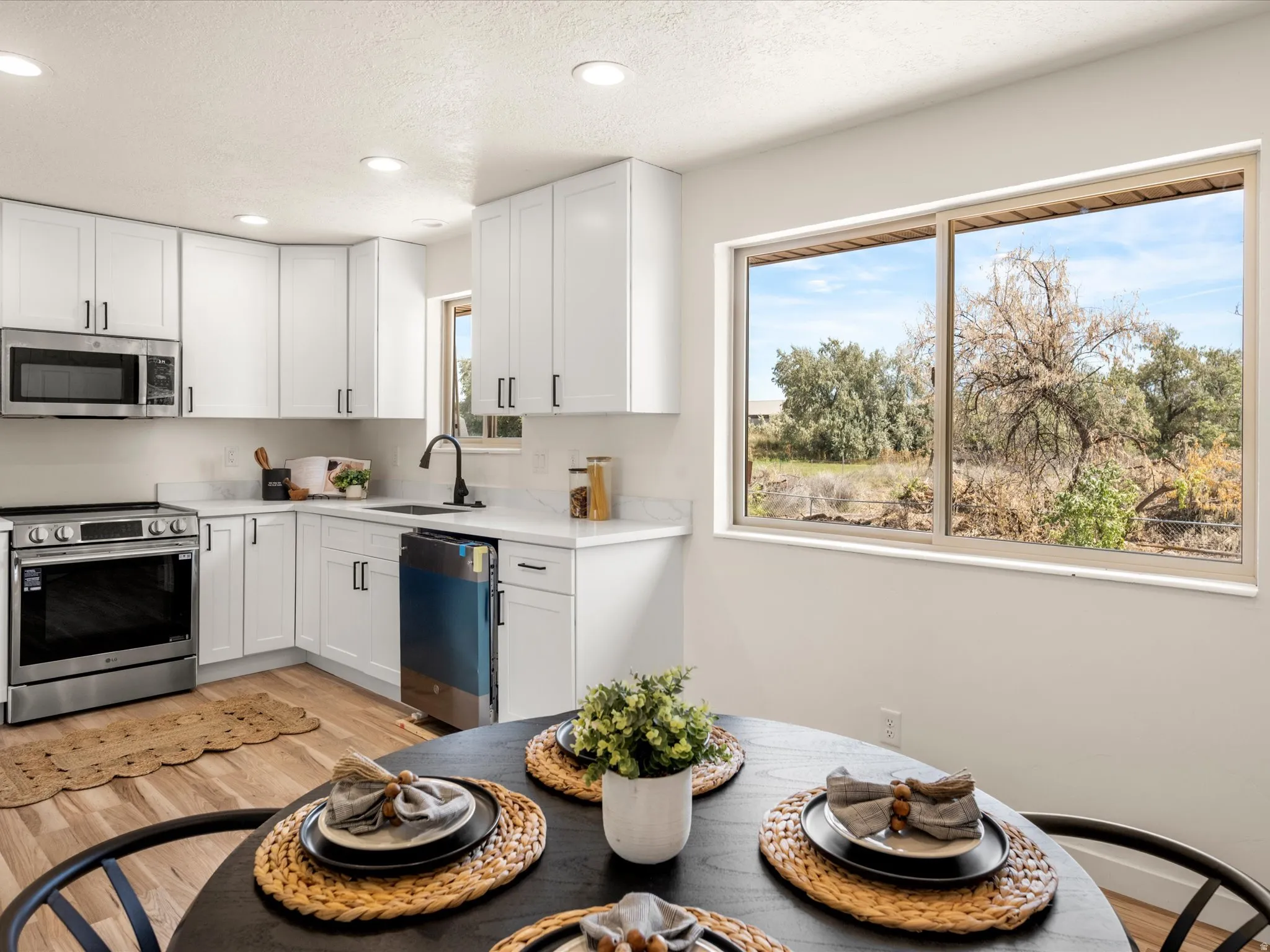 White cabinetry with dark hardware, stainless steel appliances, quartz countertops, wood-finish flooring, and a large kitchen window