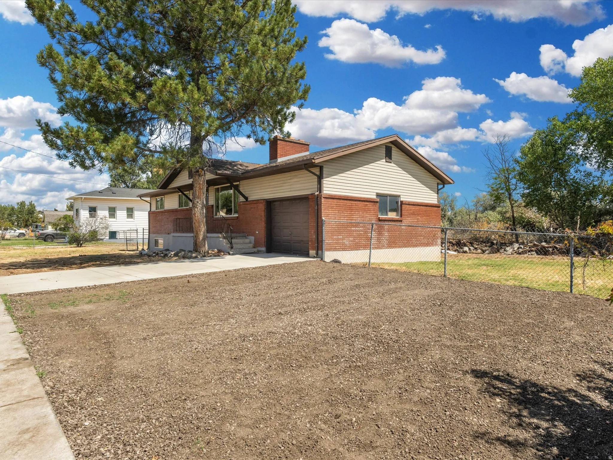 Brick and siding exterior with an attached garage, featuring a mature evergreen tree, concrete driveway, and chain-link fencing