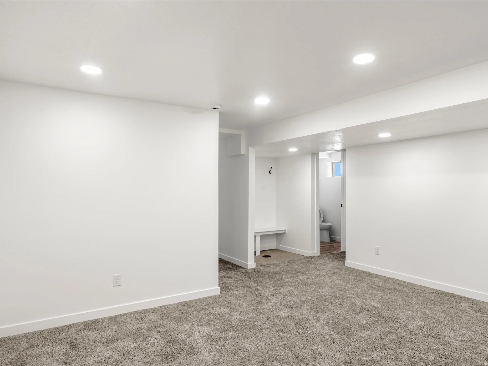 Recessed ceiling lighting, white walls, gray carpet flooring, built-in storage bench, and a partial bathroom with a toilet