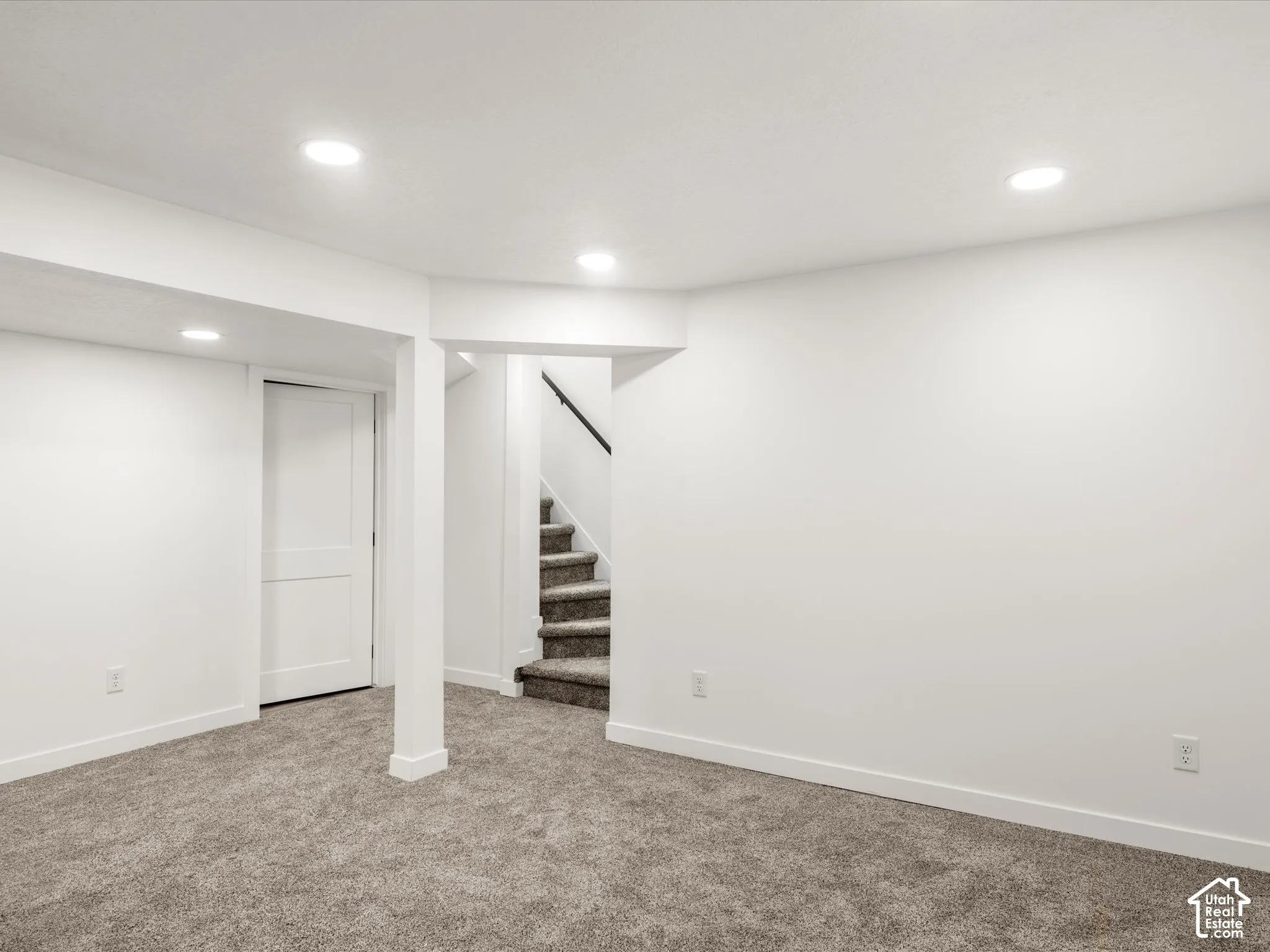 Basement family room. Laundry and cold storage on sides. Recessed ceiling lighting, white walls, gray carpeted flooring, white trim, and a staircase with a black handrail