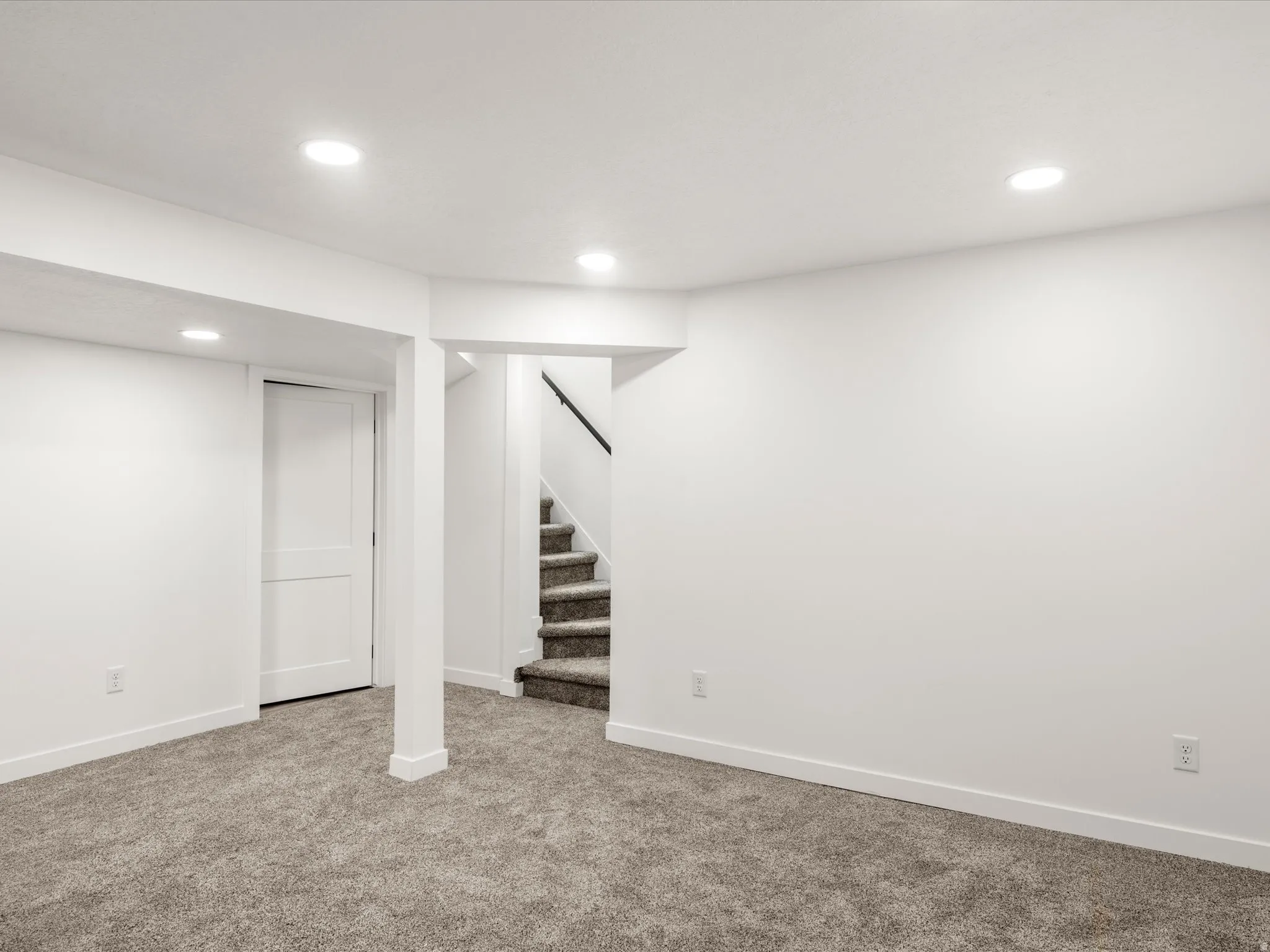 Basement family room. Laundry and cold storage on sides. Recessed ceiling lighting, white walls, gray carpeted flooring, white trim, and a staircase with a black handrail