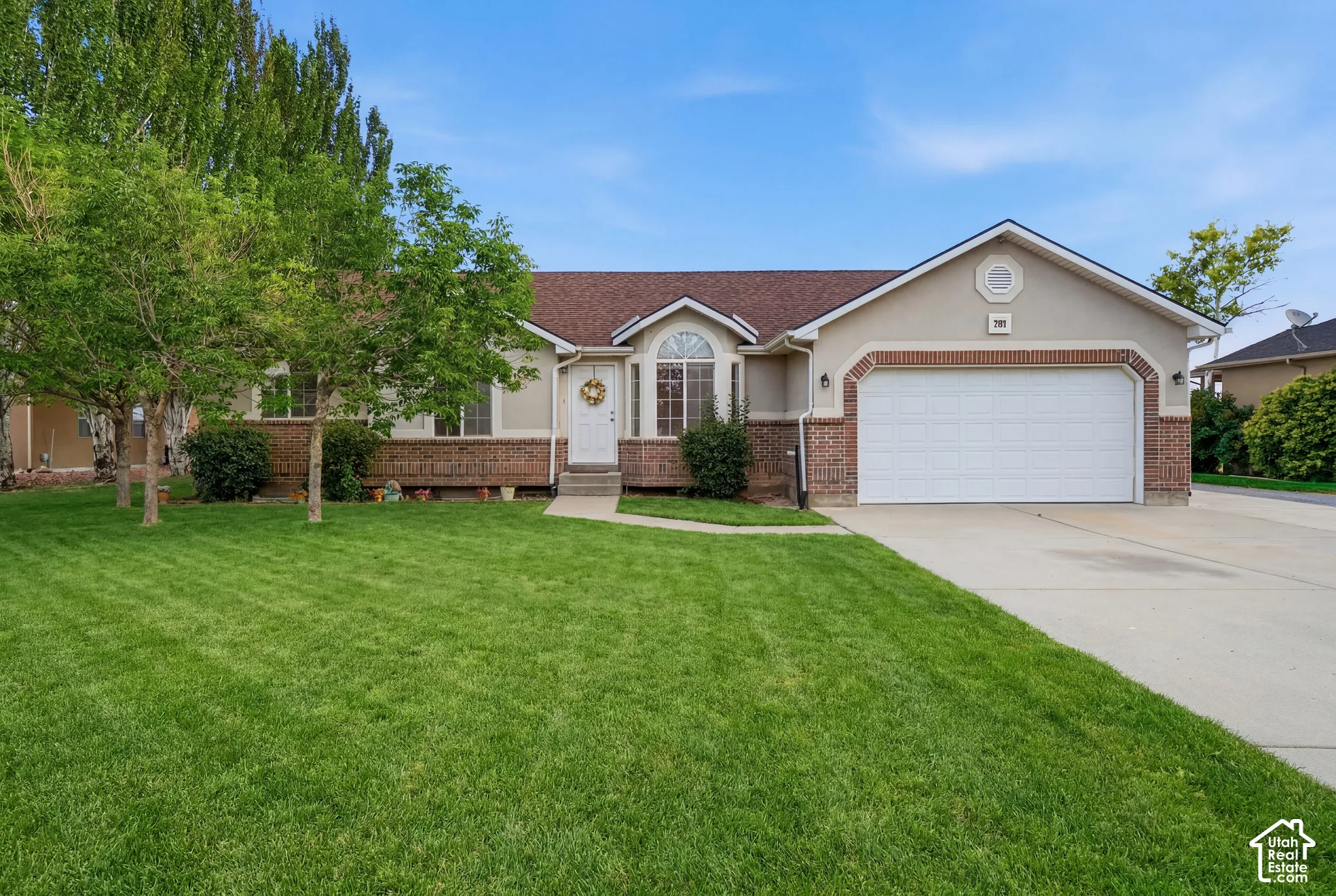 Stucco exterior with brick accents, arched window, attached garage, concrete driveway, and established front landscaping