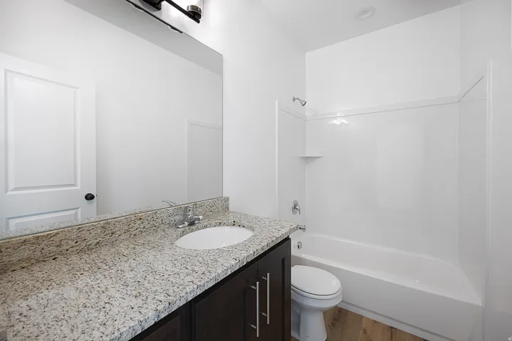 Bathroom featuring a vanity with a light-toned countertop, integrated sink, wood-finish cabinetry, a full-width mirror, and a white tub with shower surround