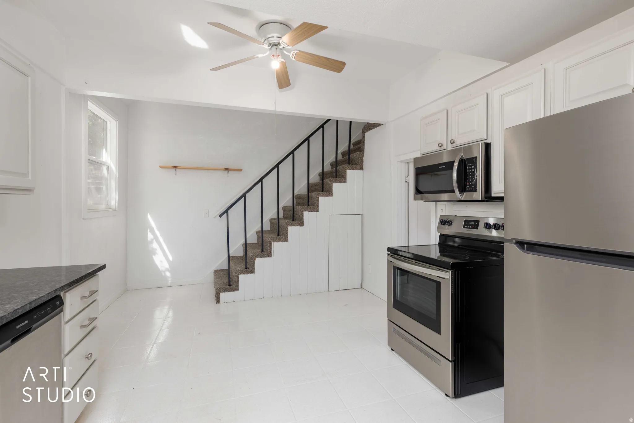 Stainless steel appliances, white cabinetry, dark stone-finish countertops, staircase with metal railing, and a ceiling fan with light fixture