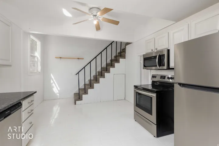Stainless steel appliances, white cabinetry, dark stone-finish countertops, staircase with metal railing, and a ceiling fan with light fixture