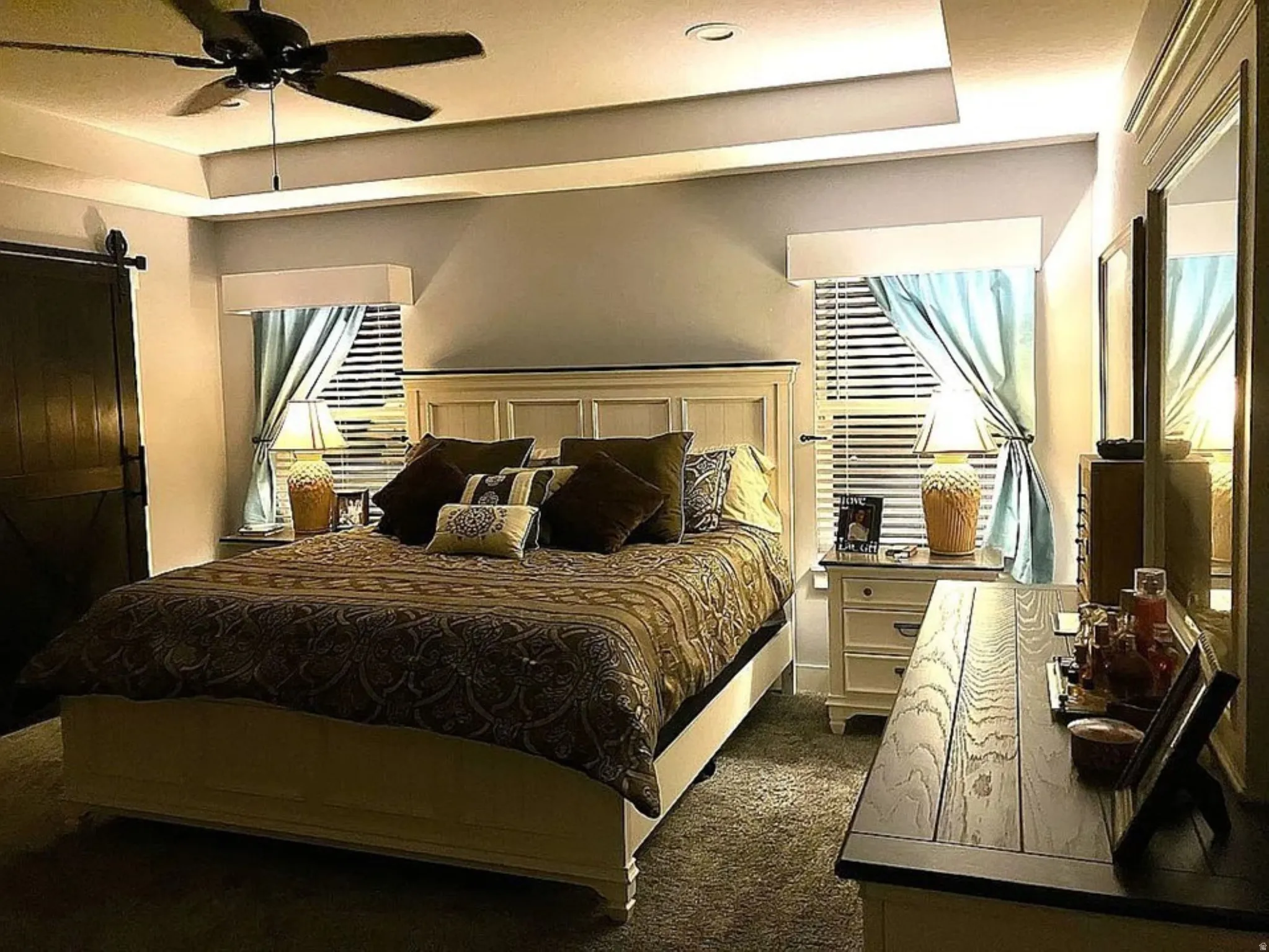 Tray ceiling with recessed lighting, ceiling fan, wood-finish barn door, white paneled bed frame, and wood-finish dresser with integrated mirror