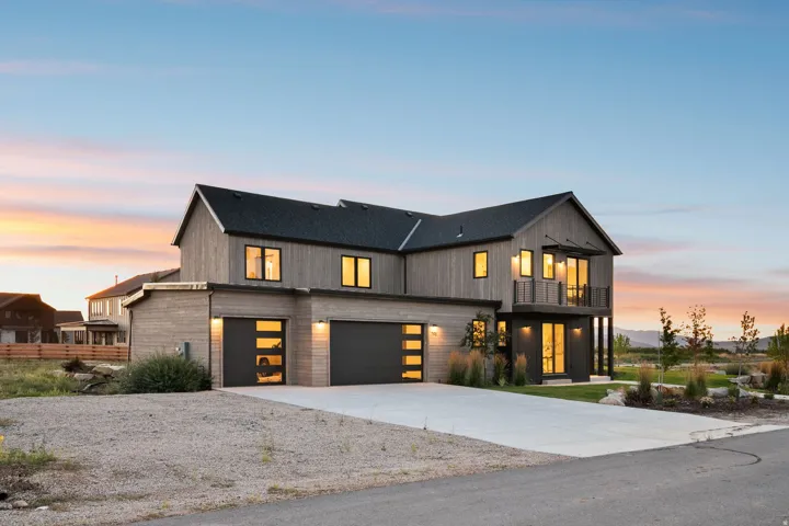 Modern exterior siding, expansive concrete driveway, dual garage doors with frosted glass panels, upper-level balcony, and surrounding landscaping