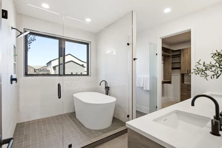 Freestanding soaking tub, glass-enclosed shower with rain showerhead, double vanity with integrated sinks, wood-finish cabinetry, and built-in closet organization