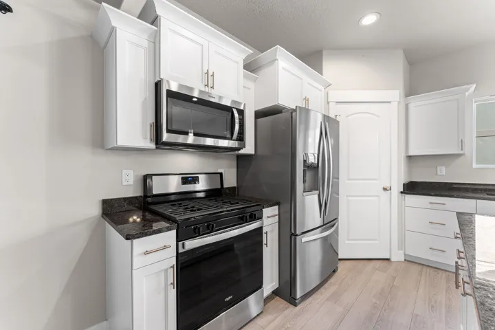 White cabinetry with gold-tone hardware, stainless steel appliances, dark stone countertops, wood-finish flooring, and recessed lighting
