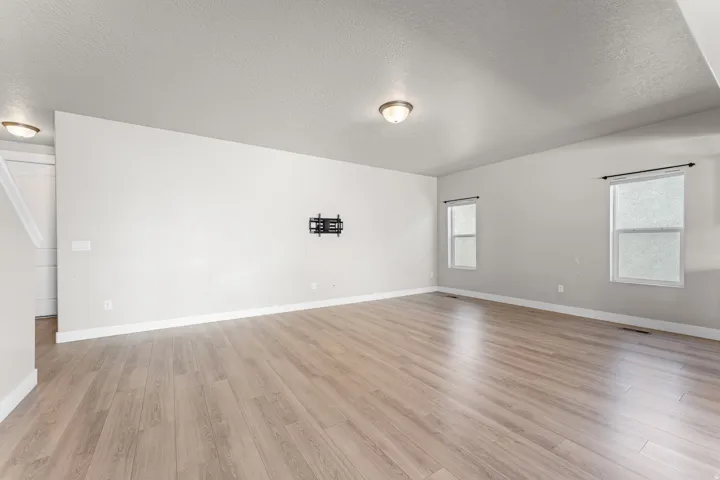 Spacious room featuring wood-finish flooring, light neutral wall tones, multiple windows with dark-finish curtain rods, and a ceiling-mounted light fixture