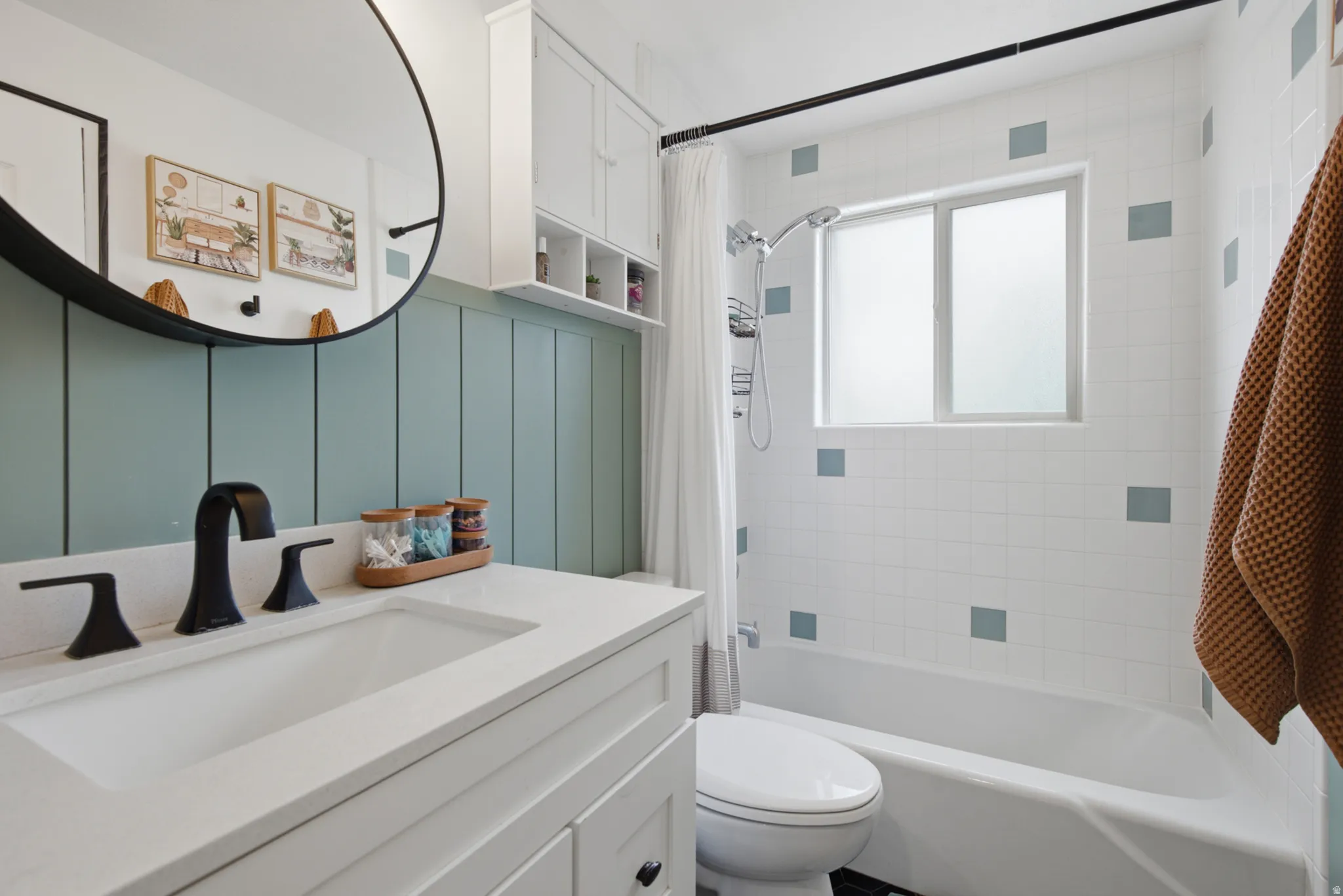 White vanity with integrated sink and matte black faucet, wall-mounted circular mirror, full bathtub with shower, and tiled walls with accent squares