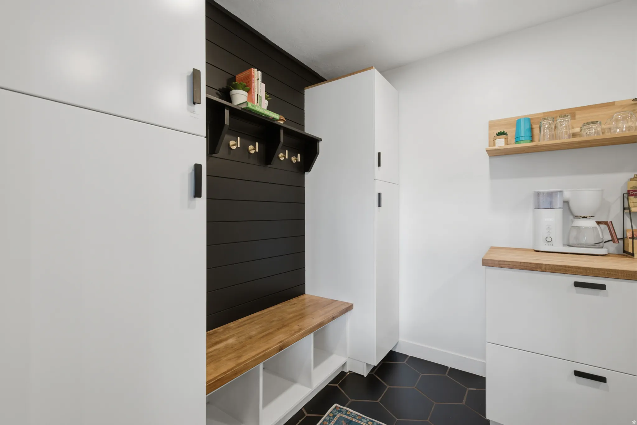 Integrated storage cabinetry with matte black hardware, black shiplap accent wall, natural wood-finish bench with cubby storage, hexagon floor tiling, and natural wood-finish floating shelves