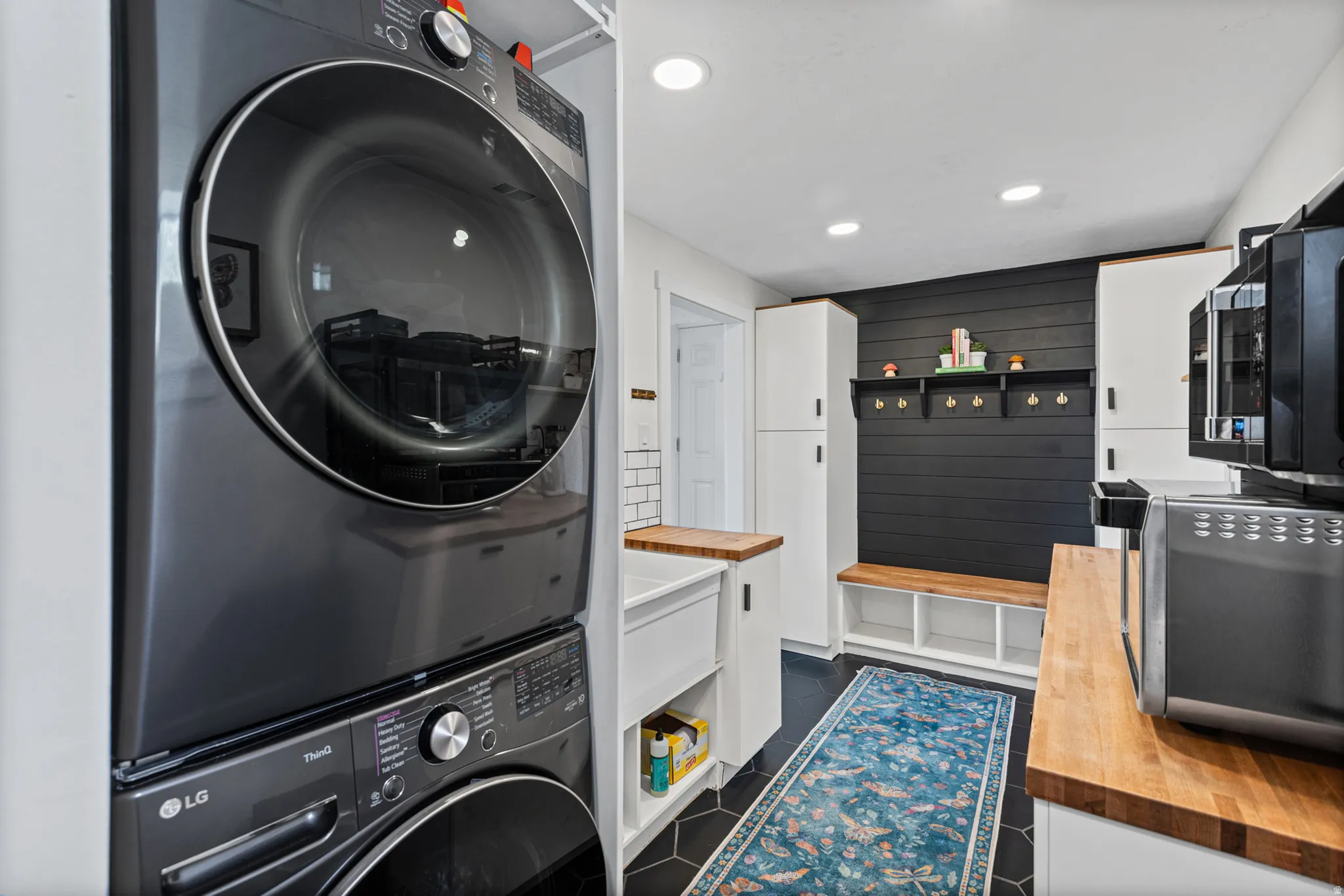 Stacked laundry appliances, built-in shelving, wood-finish countertops, black shiplap accent wall, and recessed lighting