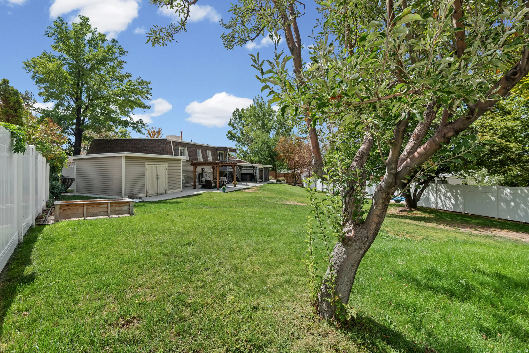 Expansive backyard featuring a manicured lawn, mature trees, a detached storage building, a concrete patio, and a white vinyl fence